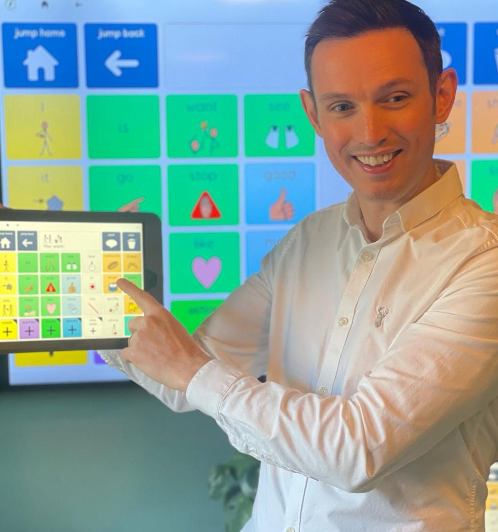 Tom McDonald smiling and pointing to an AAC device displaying a symbol grid, with a matching large display in the background