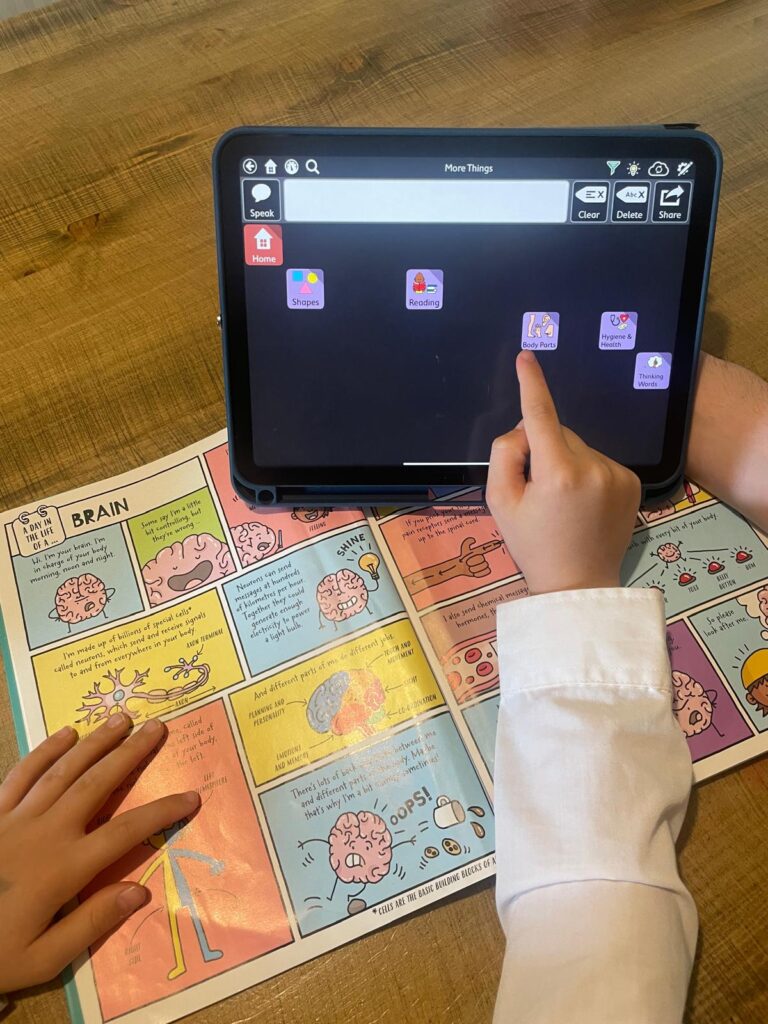 Child using an AAC device with a Grid vocabulary set alongside a colourful comic-style educational book about the brain. One hand is pointing to "Body Parts" on the device, while the other rests on the page.