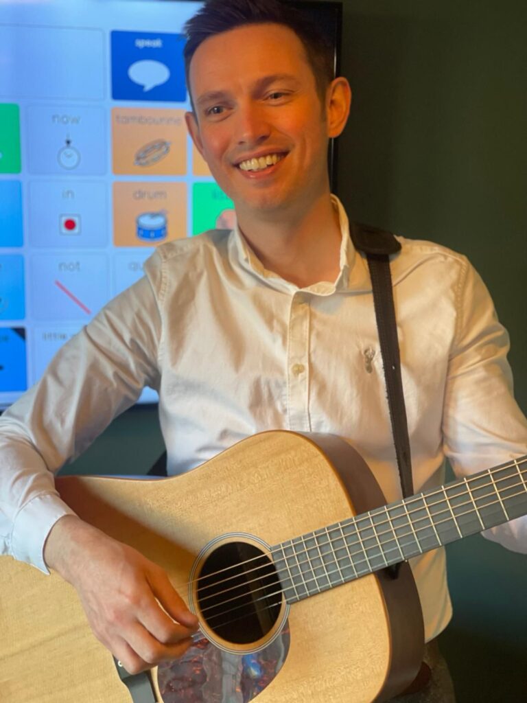 Tom McDonald smiling while playing an acoustic guitar, with an AAC symbol grid visible on a screen in the background