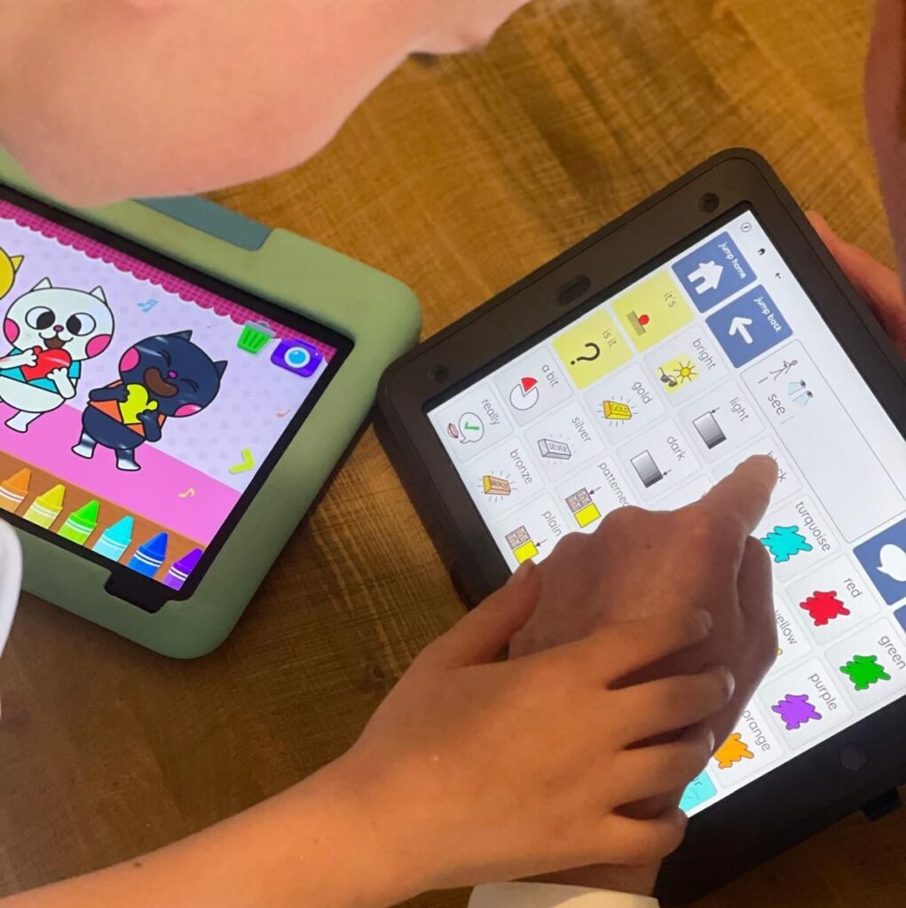 Child and adult using an AAC device together while a colourful game runs on a second tablet nearby