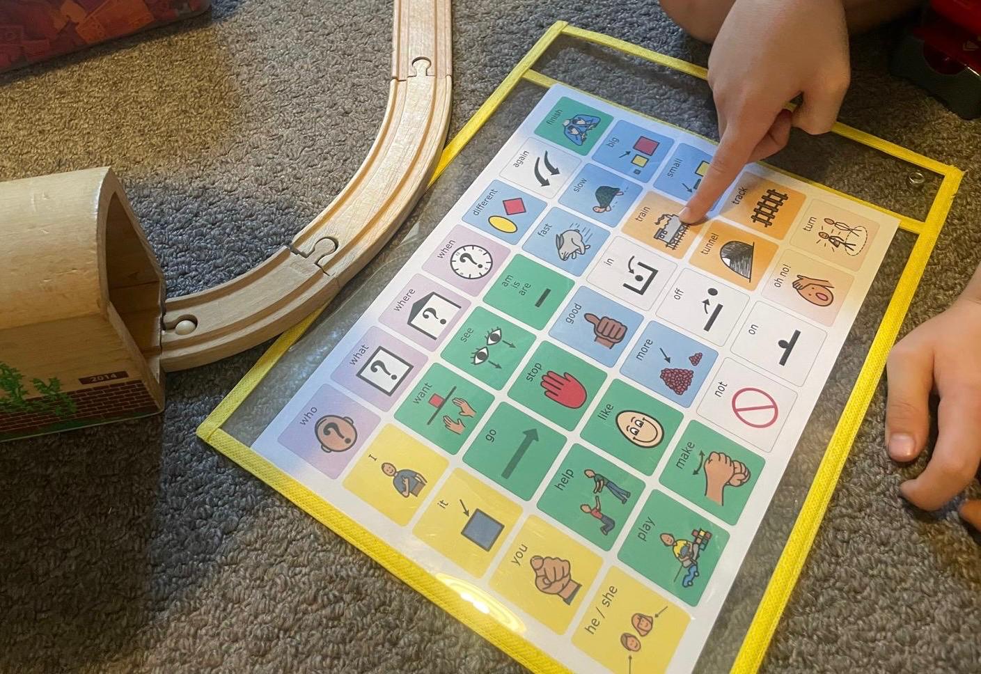 Child pointing to a laminated AAC core board while playing with a wooden toy train track