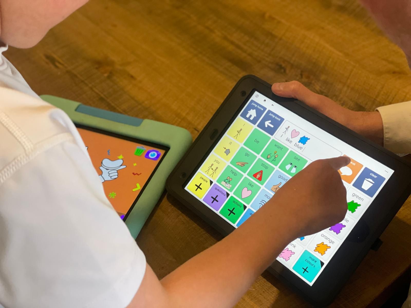 A child uses an AAC device to select the word “blue” from a colour grid, supported by an adult. Another AAC device with a cartoon thumbs-up is visible nearby on a wooden table.