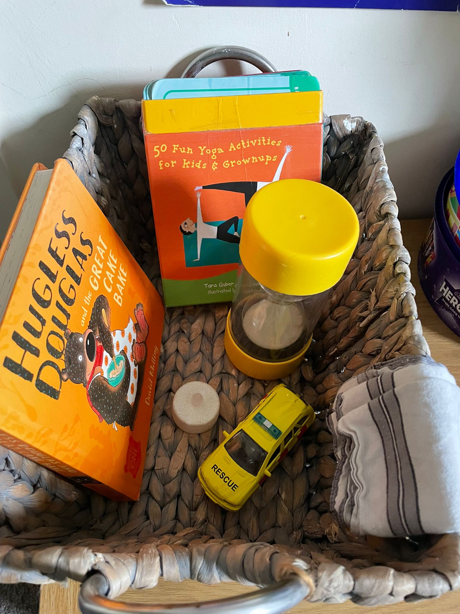 Woven basket holding a Hugless Douglas book, “50 Fun Yoga Activities” cards, a yellow sand timer, a battery tea light, a yellow rescue toy car and a folded cloth.