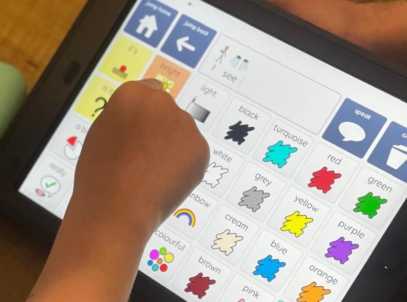 A child uses an AAC device to select the word “blue” from a colour grid, supported by an adult. Another AAC device with a cartoon thumbs-up is visible nearby on a wooden table.