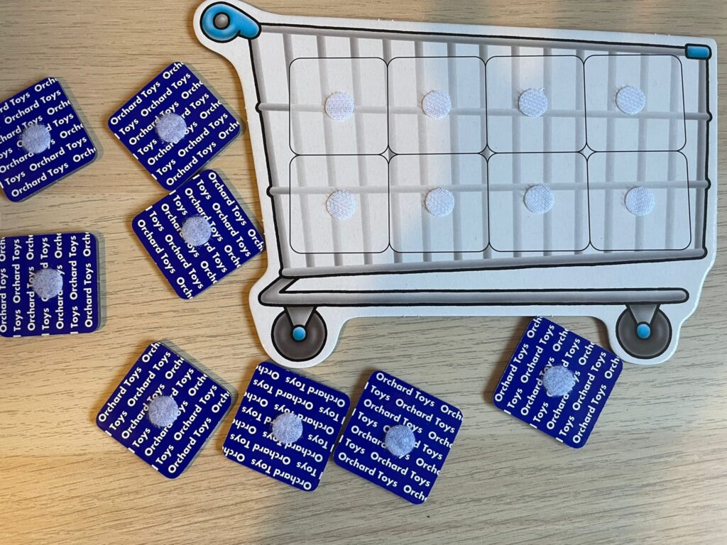A close-up photo of an adapted Orchard Toys Shopping List trolley board with Velcro dots added to each space, surrounded by the game’s blue-backed picture cards. The Velcro allows cards to stick to the trolley so items can be displayed clearly during play and viewed easily from Lottie’s eye-gaze setup.