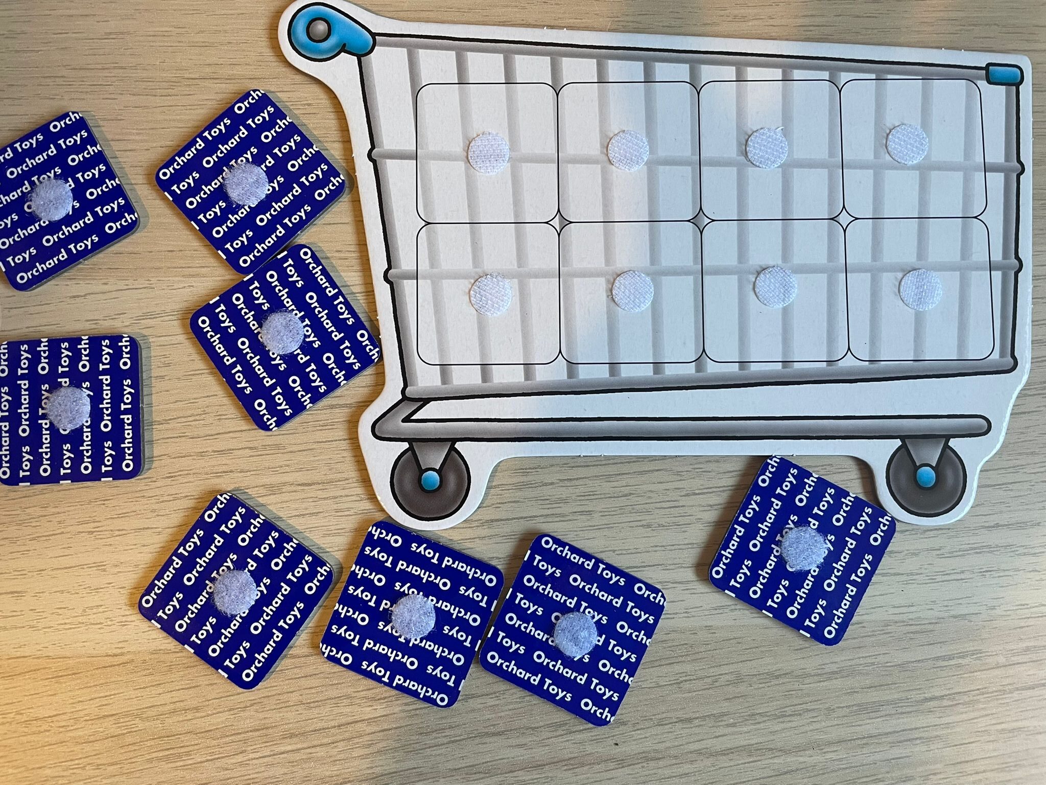 A close-up photo of an adapted Orchard Toys Shopping List trolley board with Velcro dots added to each space, surrounded by the game’s blue-backed picture cards. The Velcro allows cards to stick to the trolley so items can be displayed clearly during play and viewed easily from Lottie’s eye-gaze setup.