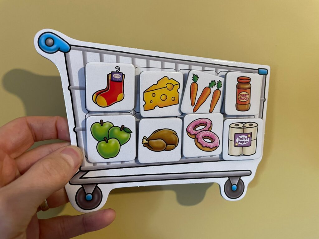 A hand holding an adapted Orchard Toys Shopping List trolley board with picture cards attached using Velcro. The cards show items such as socks, cheese, carrots, curry sauce, apples, chicken, doughnuts and toilet roll. The adapted setup helps Lottie match items from her AAC grid during the shopping game.