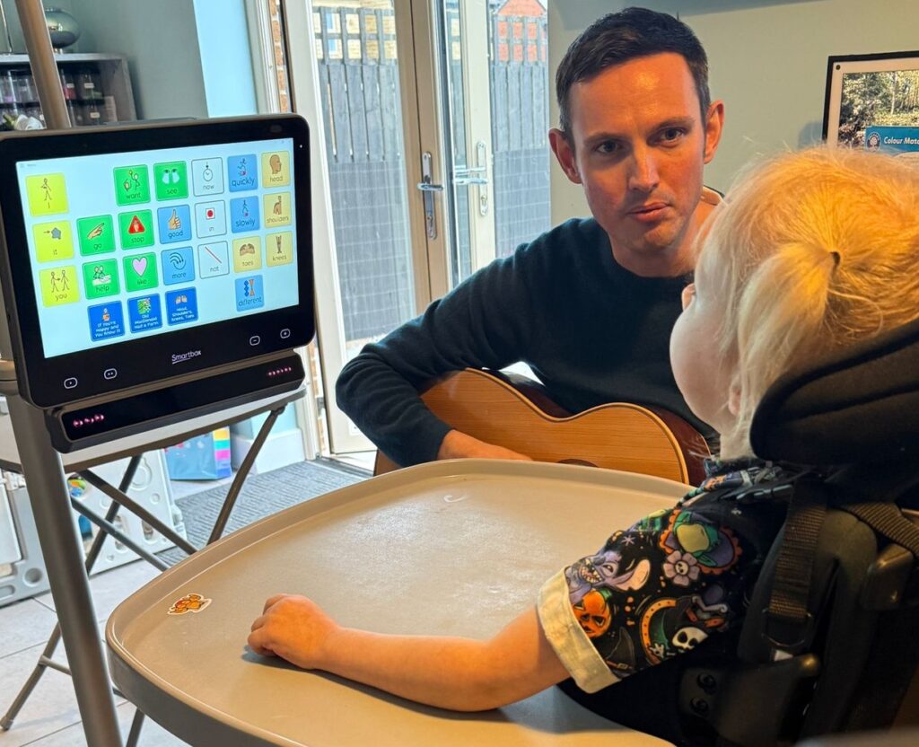 A young girl using an eye-gaze communication device with a 20-cell AAC grid displayed on the screen. She is seated in her wheelchair at a tray table, looking at the device. Across from her, an adult plays a guitar and engages her in a music activity. The session focuses on combining AAC and music to encourage communication and choice-making.