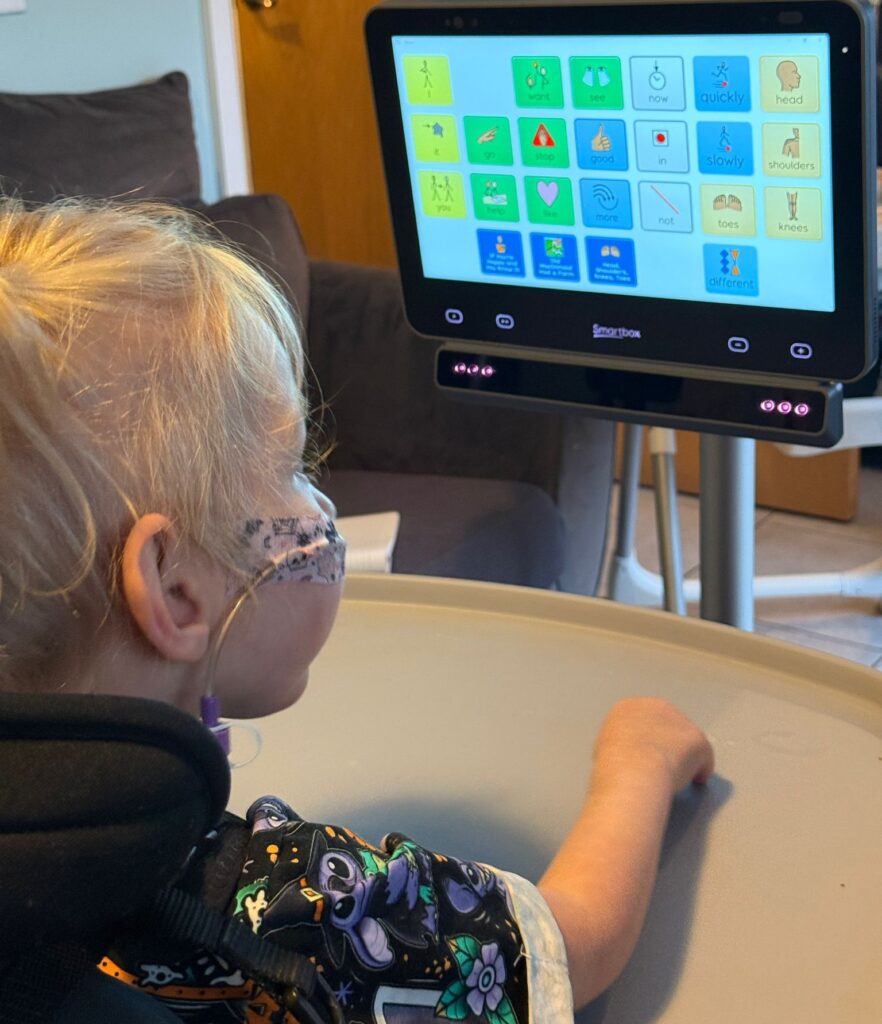 Lottie sitting in her wheelchair using her Grid Pad 13 eye-gaze communication device. The screen shows a colourful vocabulary grid with words such as “want”, “see”, “stop”, and “good”. She looks at the screen to make selections during a music activity, developing her communication skills through AAC and play.